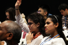 People in an audience listening to someone not visible presenting, two women are in the focus, one is raising her hand to ask a question