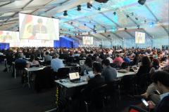 Big room with tables and people listening to a speech projected on screens